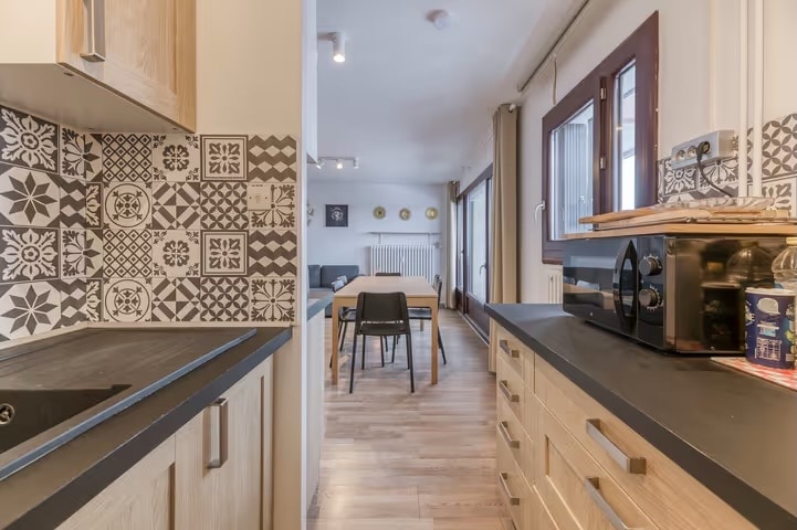 The kitchen features modern cabinetry with patterned tiled backsplash and is equipped with an induction cooktop and microwave. A view into the dining area reveals a wooden table with seating for four. Natural light streams in through large windows, enhancing the open feel.