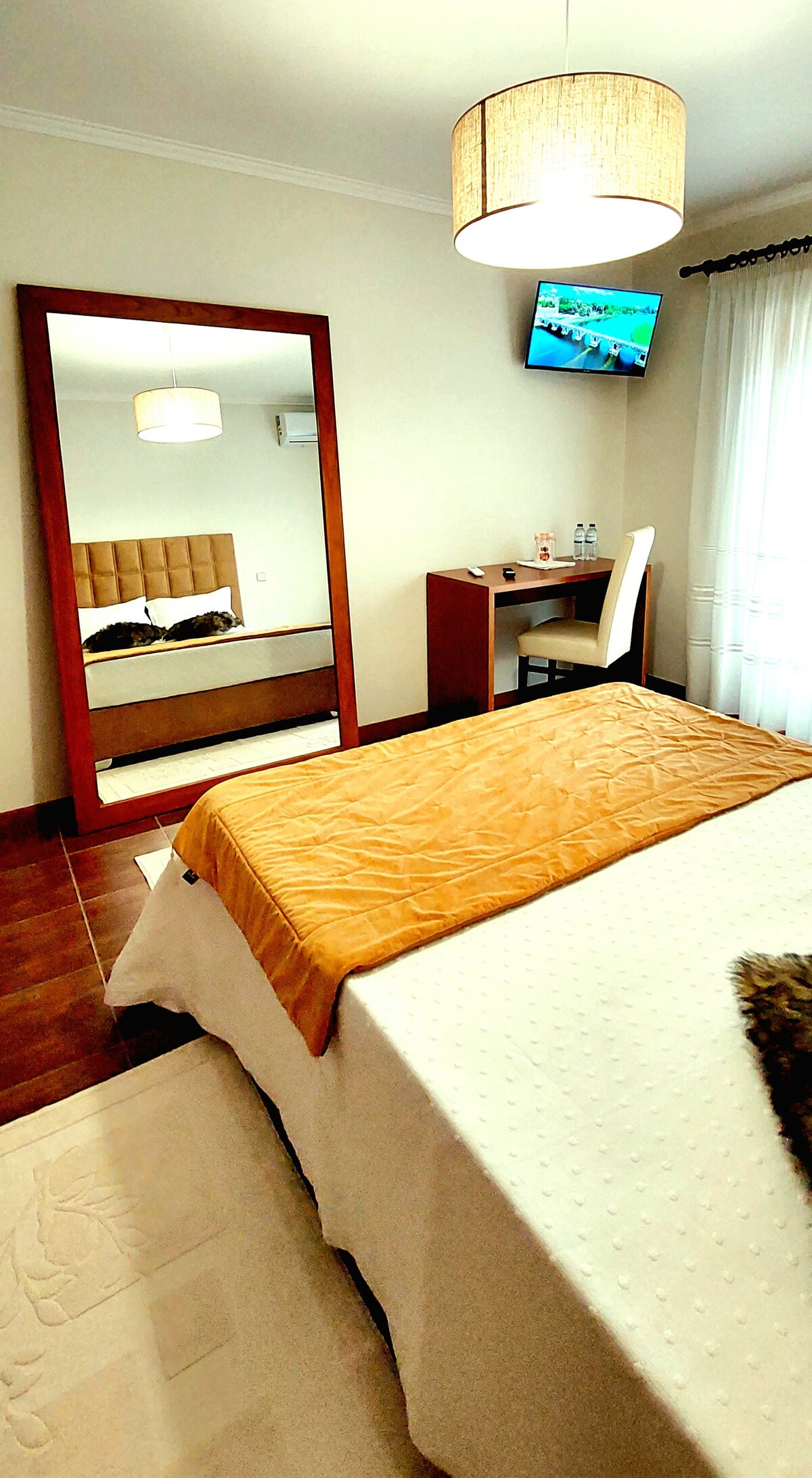 A comfortable bedroom is presented with a spacious bed featuring a light-colored blanket. A large mirror reflects the room, which includes a small desk and chair. A television is mounted on the wall. Natural light filters through sheer curtains, enhancing the inviting ambiance.