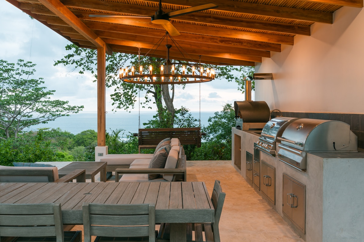 An outdoor kitchen area is highlighted by a large dining table and comfortable seating. A modern grill and pizza oven are positioned against a wall, with ocean views visible beyond the wood-beamed ceiling and decorative chandelier. A swing bench adds a relaxed touch.