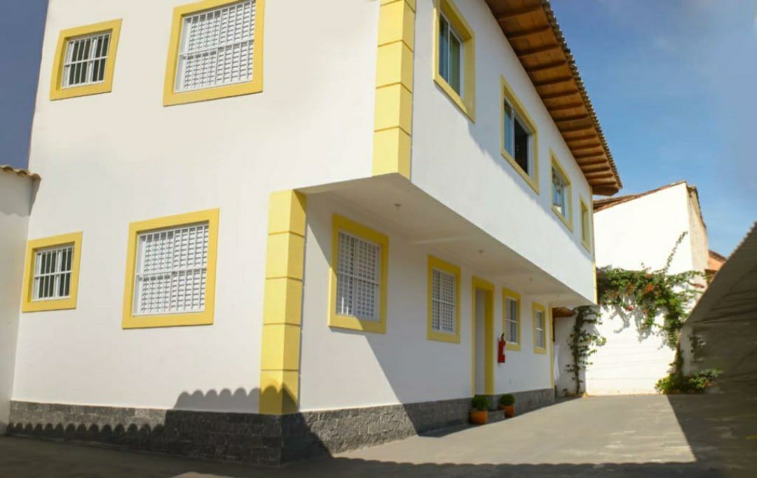 The exterior of a two-story building is shown, featuring a bright white façade with yellow accents around the windows. Multiple windows allow natural light in, while the surrounding area is neatly maintained with plants placed near the entrance.