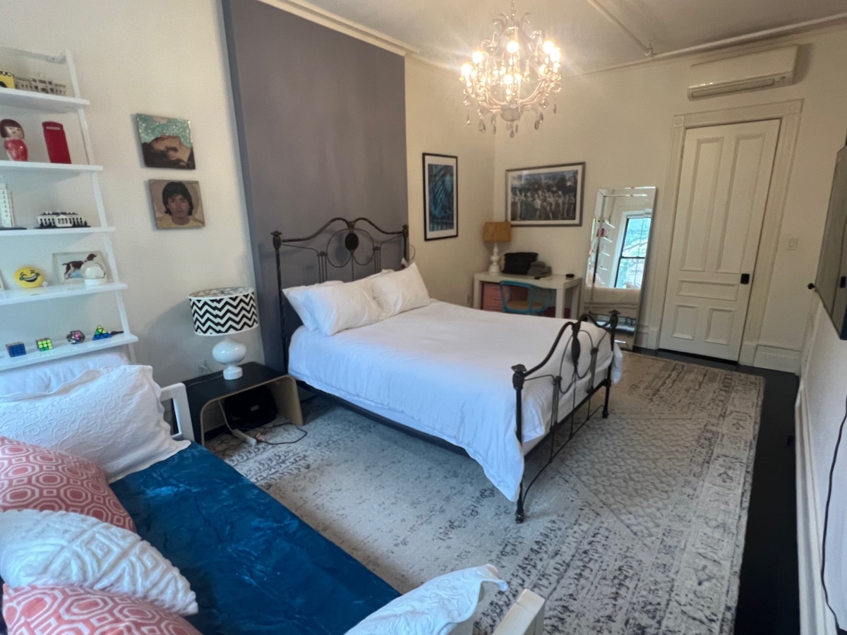 A spacious bedroom is furnished with a queen bed and a couch/daybed, complemented by a chandelier overhead. A cozy rug anchors the room, and a large window allows natural light to brighten the space. Shelving with decorative items adds a personalized touch.