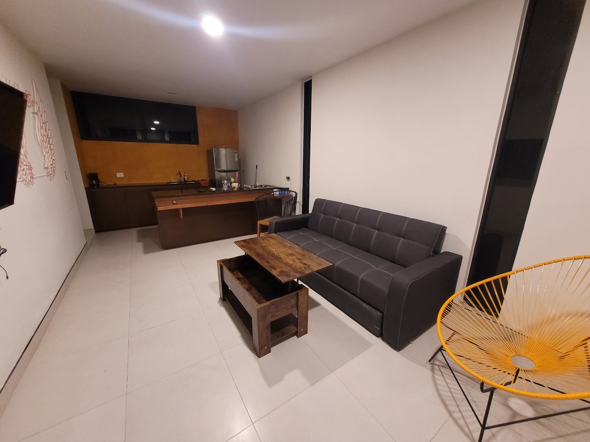 A modern living area features a sleek gray sofa positioned across from a wooden coffee table. An open kitchenette is visible with dark cabinetry, and a warm-toned wall adds contrast. A yellow accent chair brings a pop of color to the minimalistic space.