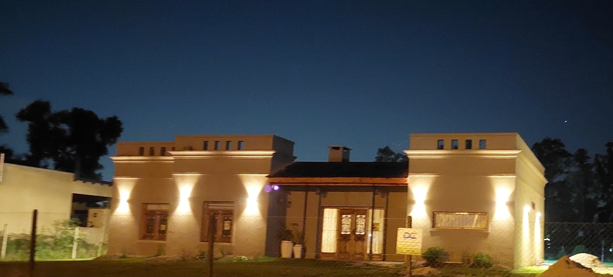 The exterior of the house is illuminated, highlighting its unique architectural features. Two symmetrical sections with decorative rooftops are visible. Soft lighting enhances the entrance, framed by tall windows, while a clear night sky creates a serene backdrop.