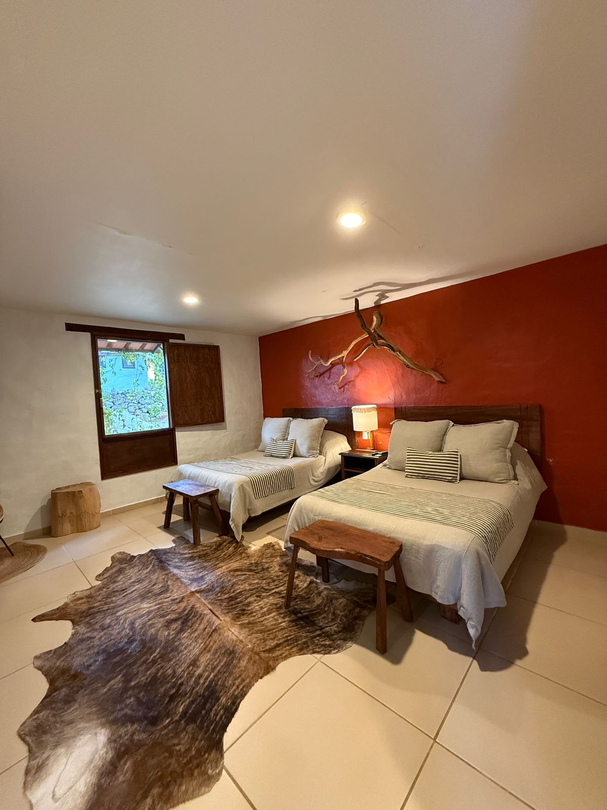 A tranquil guest room is equipped with two beds, each adorned with soft bedding and decorative cushions. A wooden lamp is placed beside a bed, while a small natural wood structure serves as a side table. A window allows natural light to brighten the space.