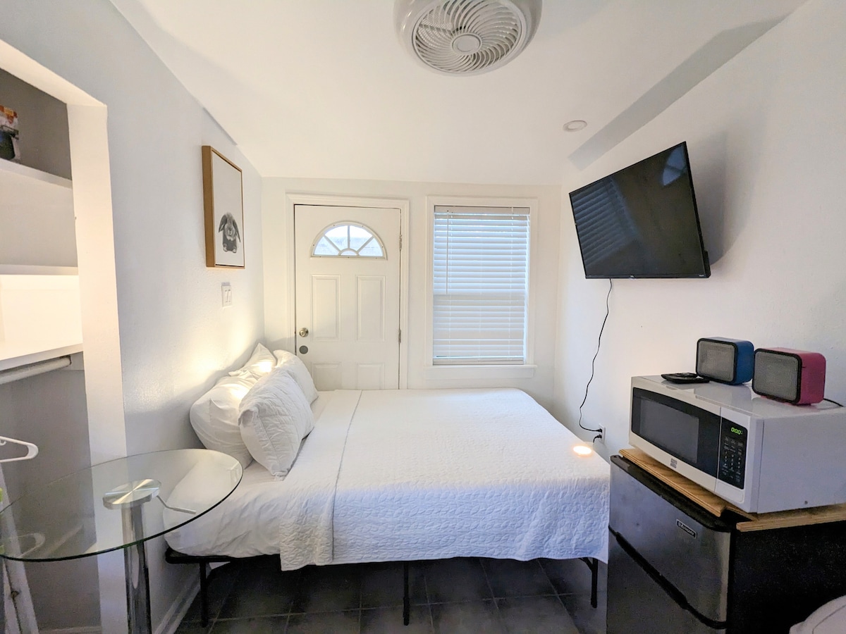 A compact bedroom features a cozy mattress topped with white bedding. A small glass-topped table sits beside the bed. A microwave and mini fridge occupy one corner, while a television is mounted on the wall opposite the bed. Natural light enters through a window near the door.