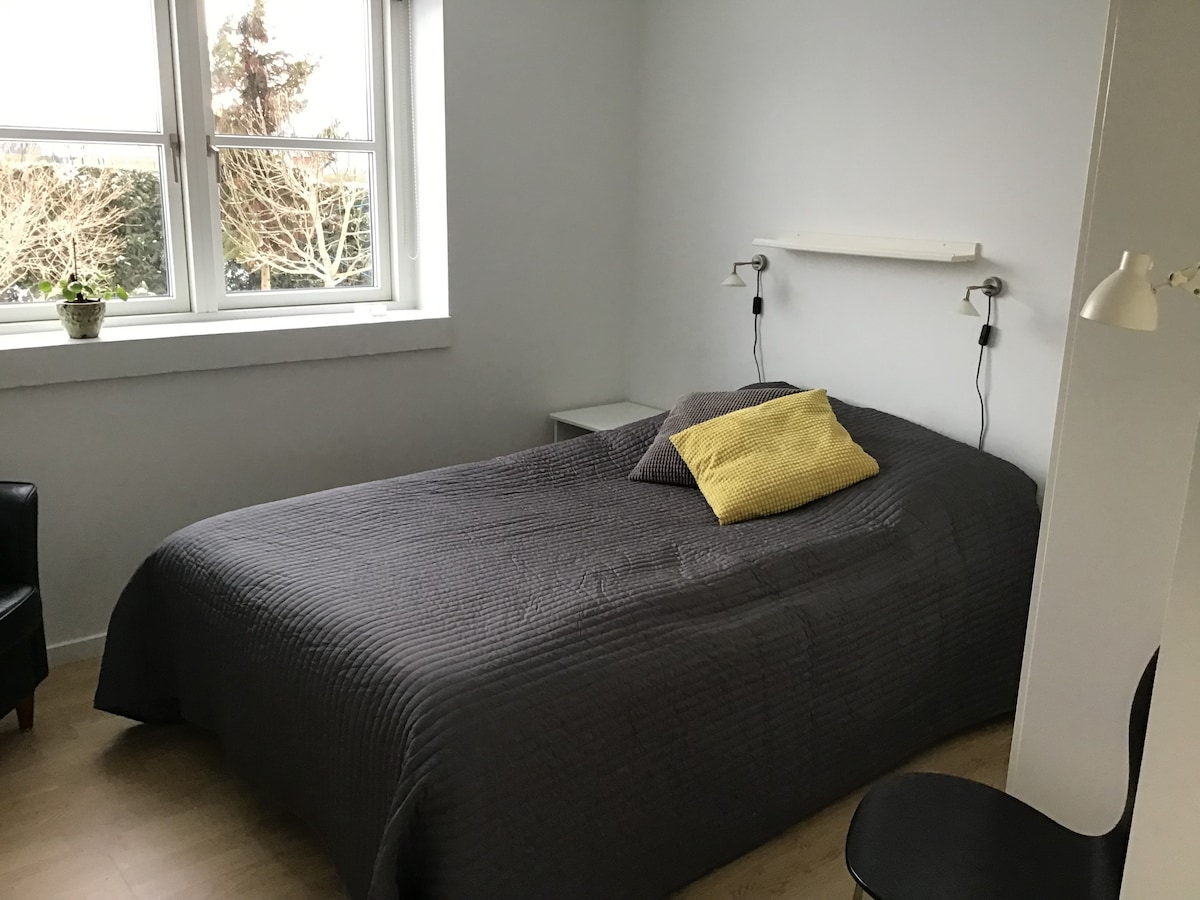 A calm bedroom features a spacious bed covered with a textured gray blanket and adorned with two decorative pillows. Natural light enters through a large window, illuminating a small side table. A reading lamp and a chair are also visible in the room.