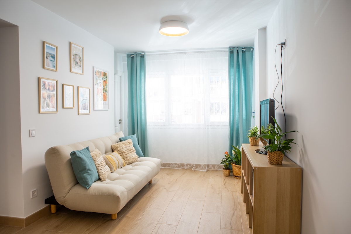 The living area features a light-colored futon adorned with decorative cushions, complemented by wooden flooring. A window with sheer curtains allows natural light to fill the space. A wooden media unit houses a television, while decorative plants add touches of greenery.