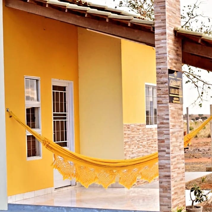 A bright yellow exterior features a comfortable hammock, inviting relaxation. The entrance is framed by two windows, allowing natural light to enhance the cheerful ambiance. The structure is complemented by natural stone accents, creating an appealing visual contrast.