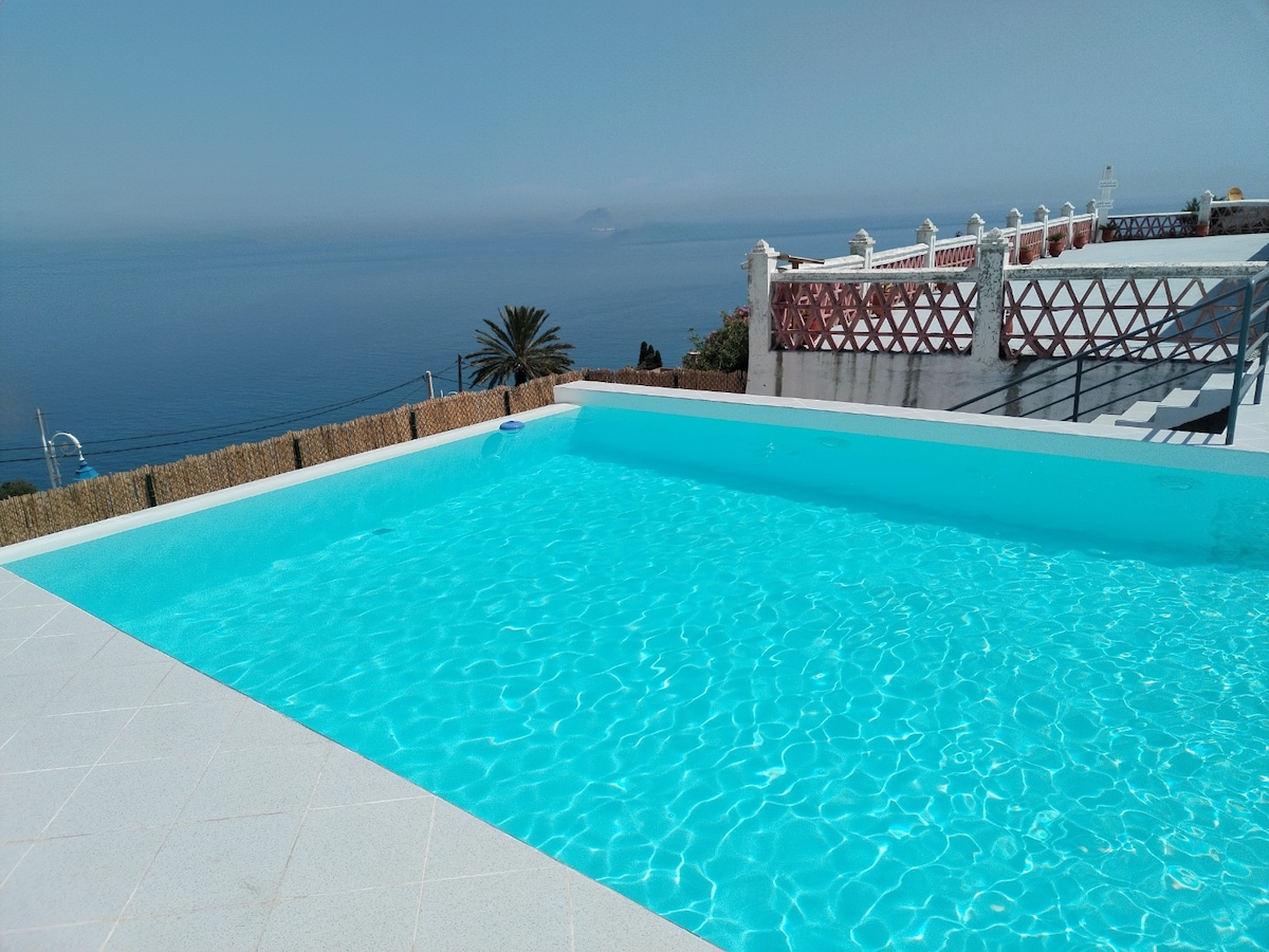 A large rectangular swimming pool is filled with clear turquoise water, reflecting the sky above. The pool area features light-colored tiles and a view of the ocean in the background, along with a terrace space bordered by a decorative fence.