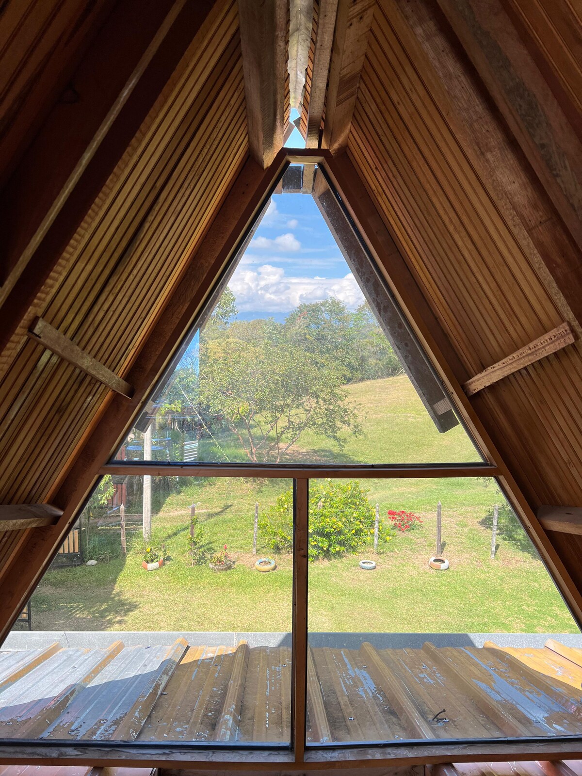 A triangular window frames a view of lush greenery and a blue sky, inviting natural light into the space. The wooden interior features exposed beams and slatted wood details, creating a cozy atmosphere.