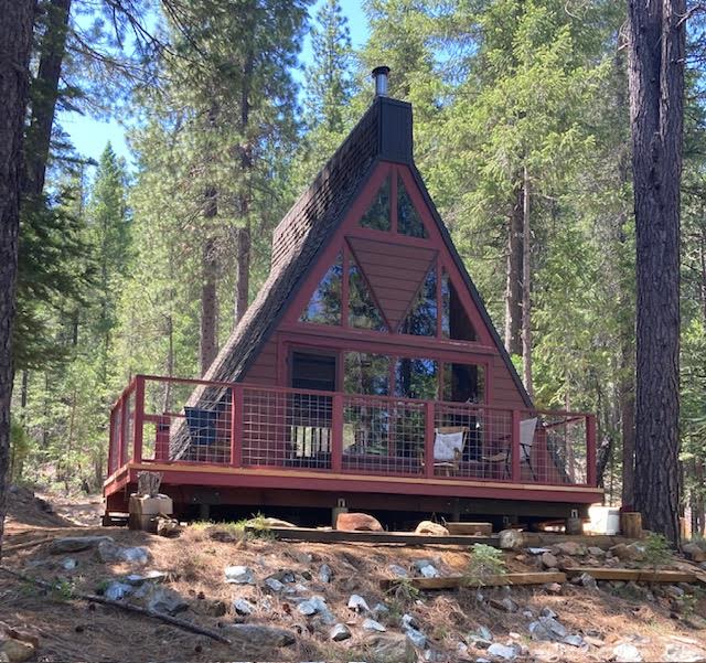 The cabin features a distinctive A-frame design, surrounded by tall trees within a forested setting. A wraparound deck offers space for outdoor seating, with large windows allowing natural light to enhance the interior. Neutral-toned materials complement the natural surroundings.