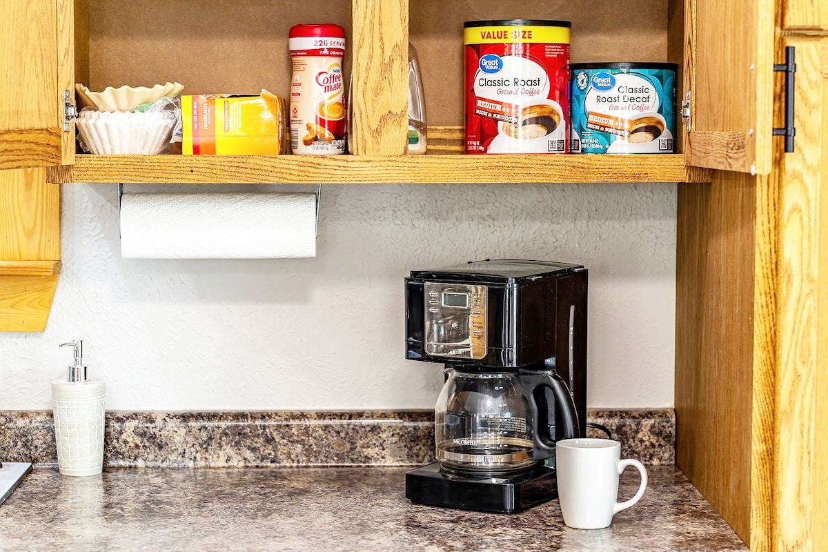 A functional kitchen space is showcased, featuring an electric coffee maker with a glass carafe. Above it, shelves are stocked with coffee supplies, including various canisters. A white mug sits on the countertop, alongside a roll of paper towels for convenience.
