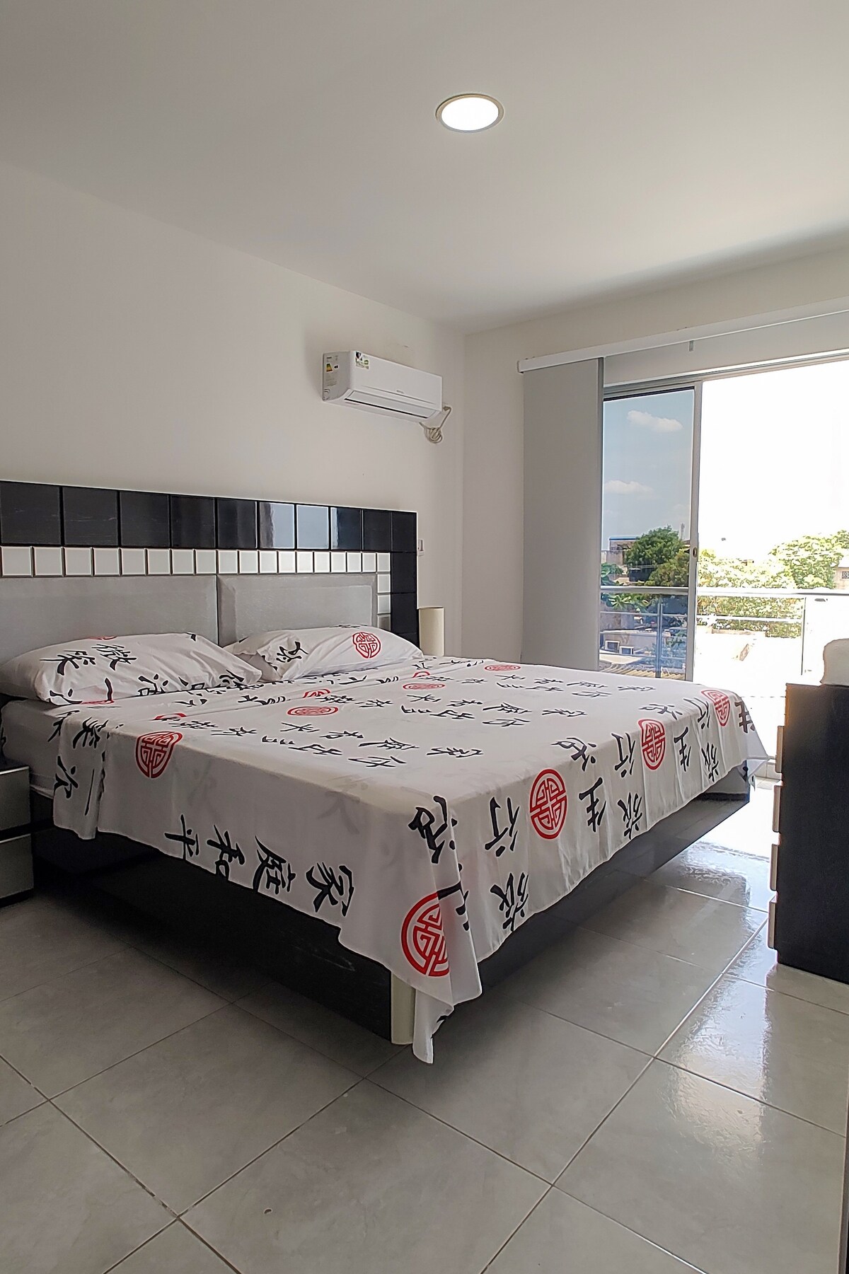 A well-lit bedroom features a spacious bed dressed in a simple patterned blanket. A large window offers natural light and views of the outdoors. Air conditioning is provided for comfort, and minimalistic bedside tables are placed beside the bed.