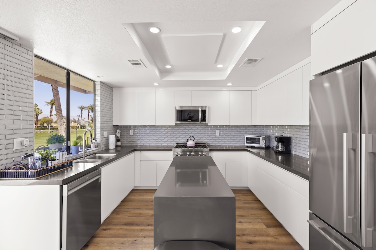 A modern kitchen is presented with sleek white cabinetry and gray countertops. A spacious island stands at the center, complemented by stainless steel appliances. Large windows allow natural light to flood the space, providing views of the outdoor scenery.