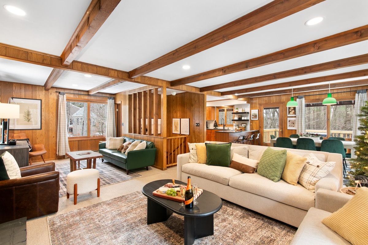 An inviting open-concept living area features wooden beams and large windows, allowing for natural light. A spacious cream sofa, accent chairs, and a round coffee table create a comfortable seating arrangement. A well-equipped kitchen is visible in the background, along with a dining area for gatherings.