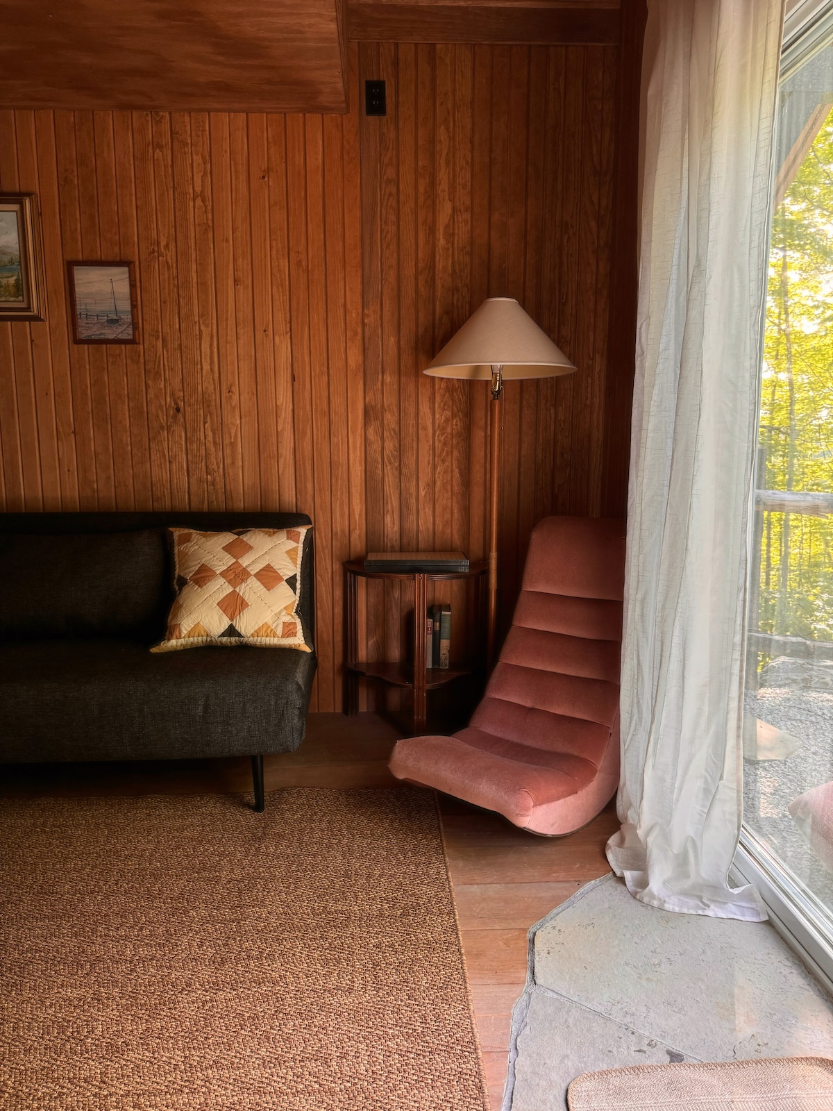 A cozy living room corner is showcased with a dark sofa and a vibrant pink accent chair. Natural light filters in through large windows, illuminating the warm wooden paneling and textured rug on the floor. A floor lamp provides additional lighting.