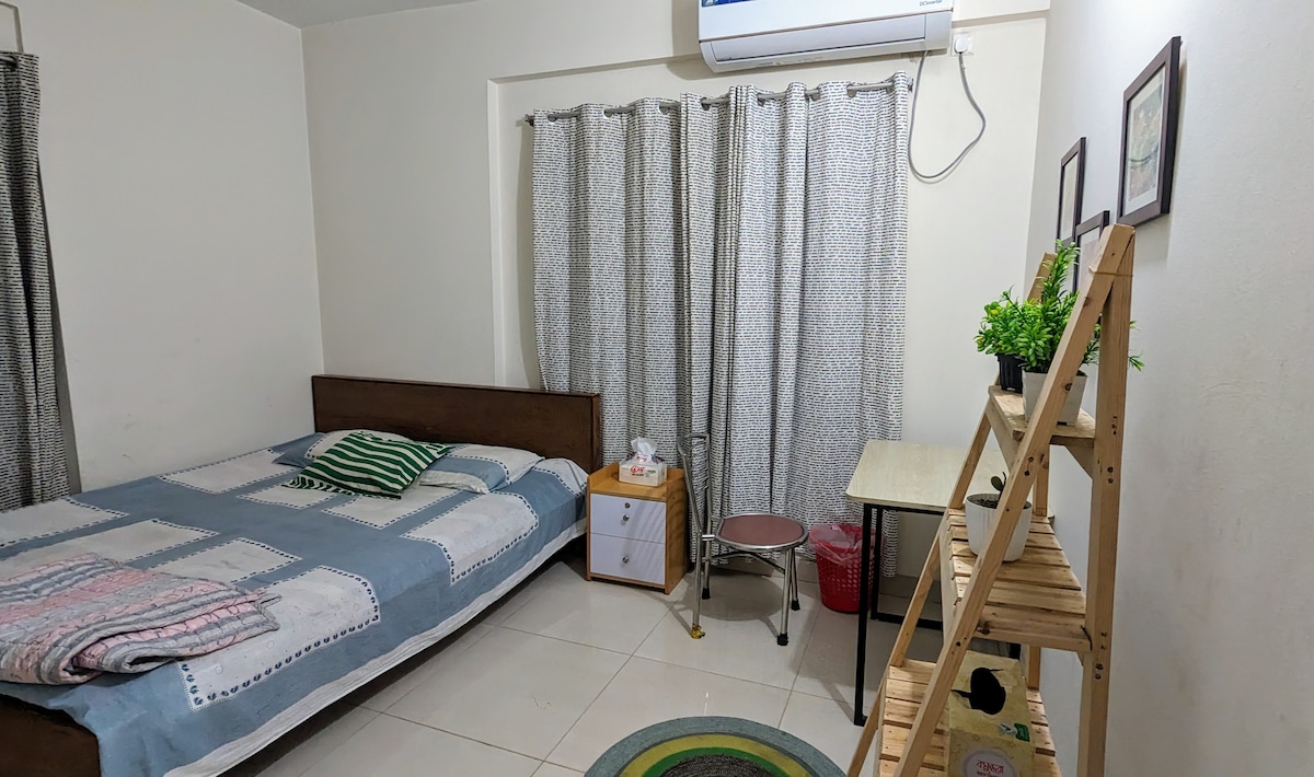 A simple bedroom features a wooden bed draped in a patterned blanket. Nearby, a small table with a chair and a decorative plant adds charm. Soft curtains frame the window, while a neatly organized shelf holds decorative items, enhancing the room's functionality.