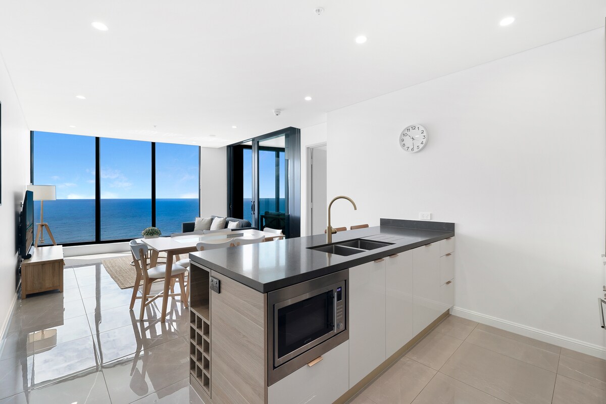 The image features a modern kitchen with a sleek design, equipped with a built-in microwave and a wine rack. A spacious living area is visible through large glass windows, showcasing stunning ocean views. Bright natural light fills the open space, enhancing the clean, contemporary aesthetics.