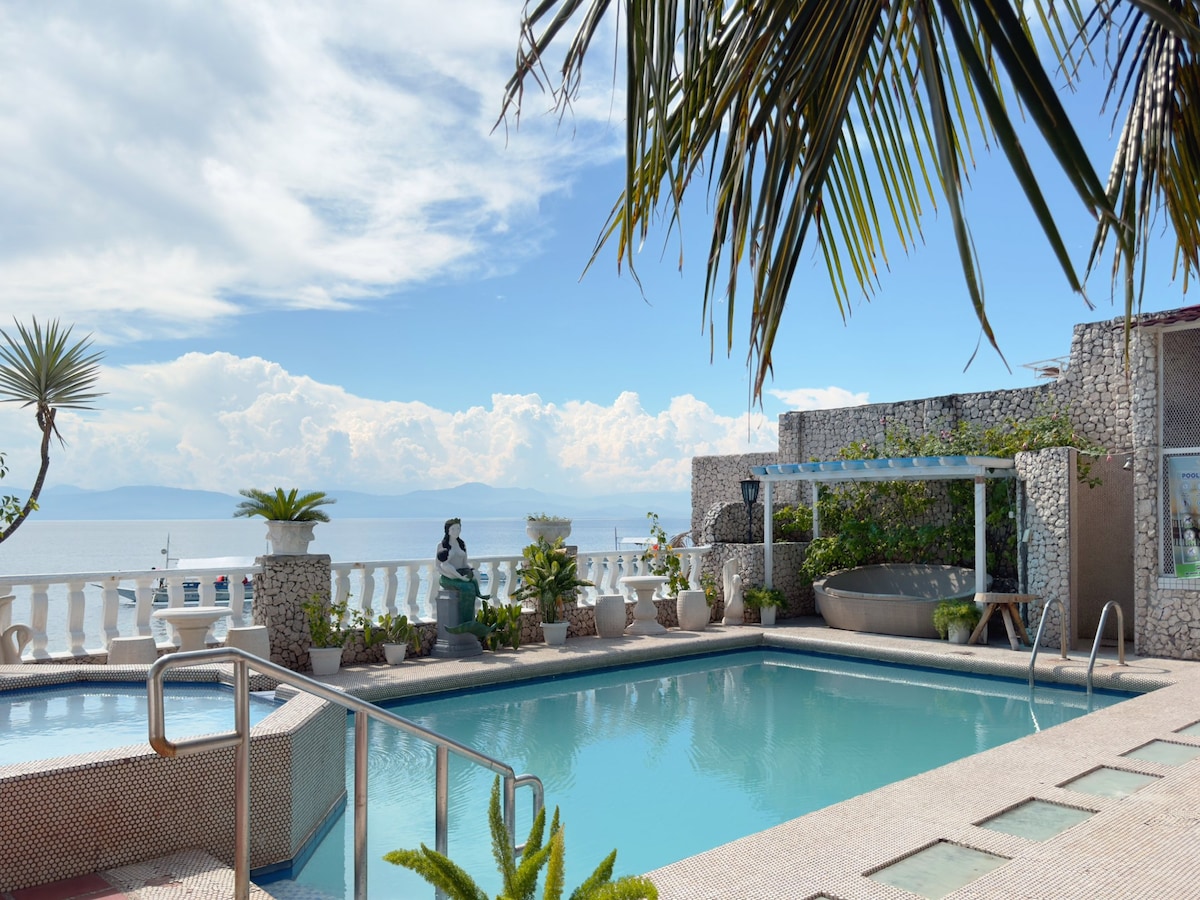 A serene pool area is framed by palm trees, with clear water inviting relaxation. The backdrop showcases the ocean and a horizon with soft clouds. Lush plants in pots and decorative statues enhance the tranquil setting, creating a peaceful retreat by the water.