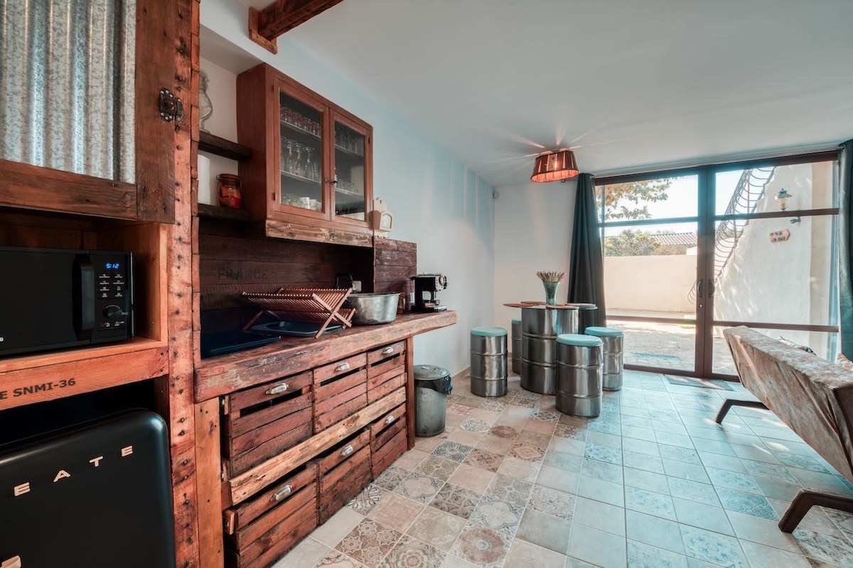 A rustic kitchen area displays wooden cabinetry and a farmhouse-style sink. Metallic stools surround a small table, while large glass doors provide access to an outdoor space. Unique flooring features a mix of tile patterns, adding character to the room.