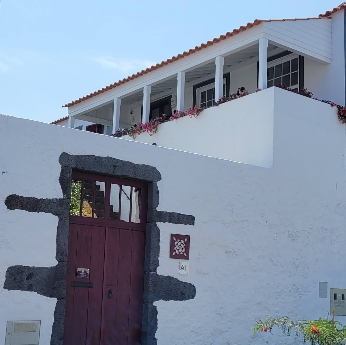 The image displays a historic stone wall accentuated by a wooden door painted in deep red. Above, a balcony showcases a white structure with large windows, adorned with flowering plants along the edge.