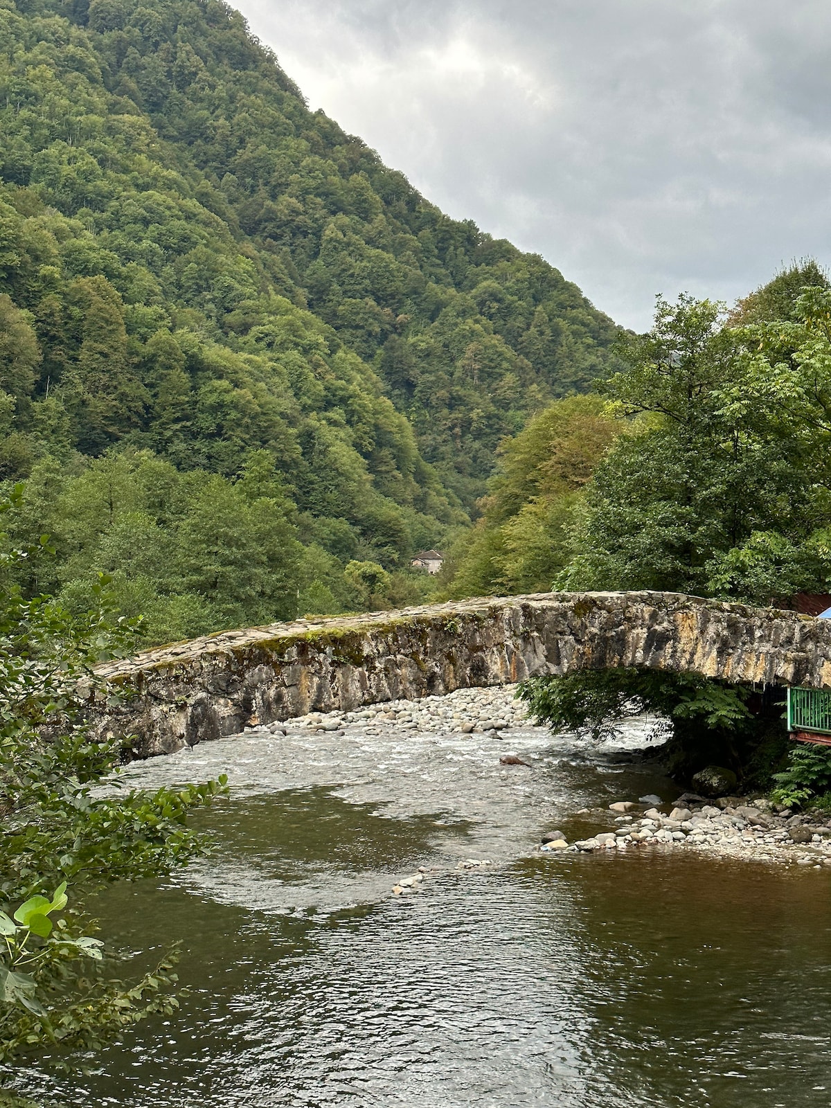 An ancient stone bridge is seen arching over a flowing river, surrounded by lush green hills. The vibrant foliage of trees creates a serene backdrop, while soft clouds drift across a grey sky, reflecting the natural beauty of the landscape.