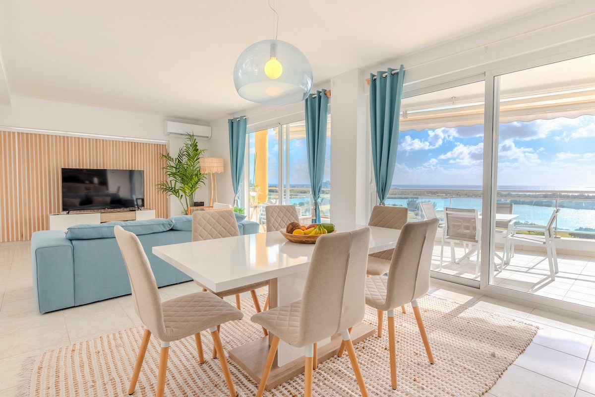A bright dining area features a white table surrounded by six light-colored chairs. Large windows provide a view of the outdoor space. A comfortable blue sofa is positioned nearby, and a modern light fixture hangs above the table, adding style to the inviting interior.