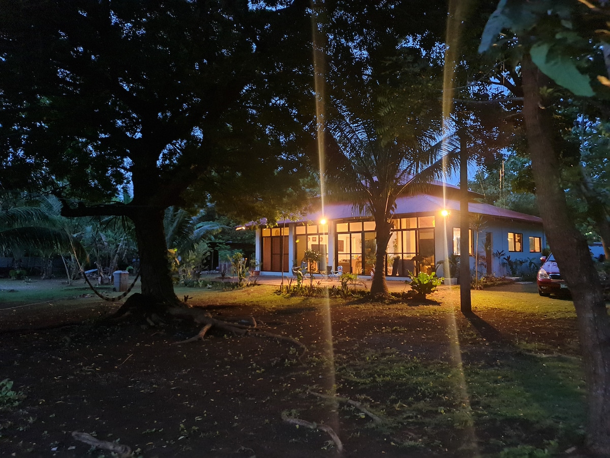 The exterior of the beachfront retreat is subtly illuminated at dusk, showcasing large glass windows that reflect warm light. Surrounding trees offer a natural canopy, while the lush landscape creates a serene environment. A vehicle is parked nearby, emphasizing accessibility.