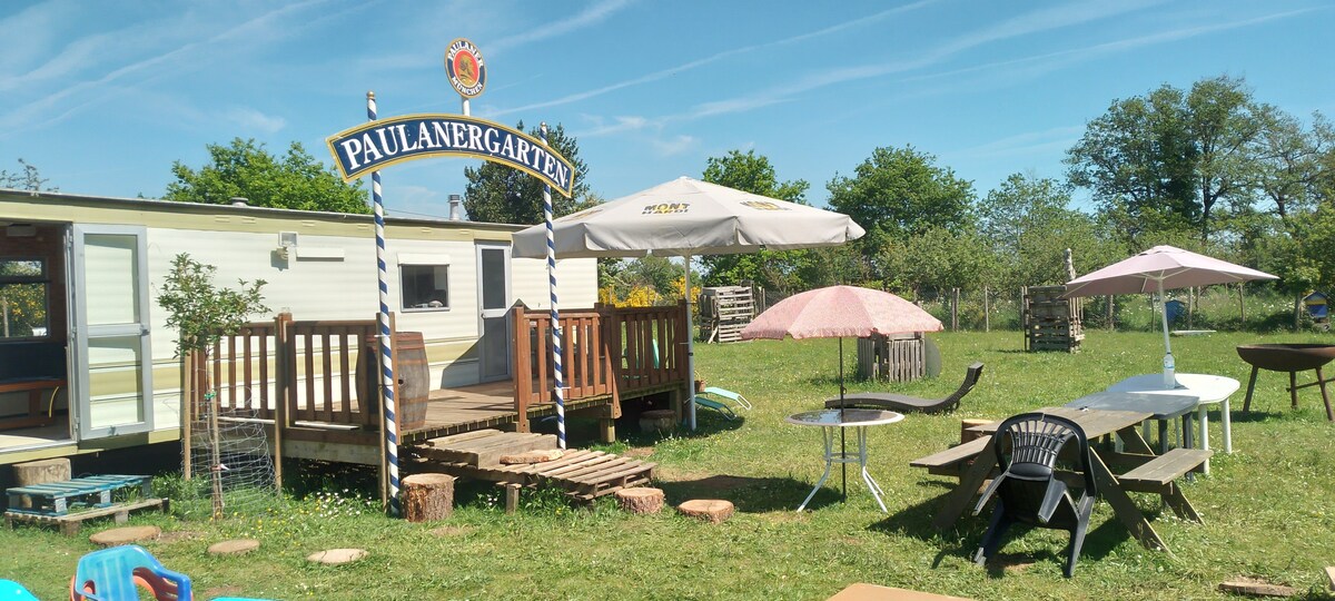 An outdoor area features a mobil home with a wooden deck, surrounded by grassy terrain. A large umbrella offers shade over a table and chairs, while additional outdoor seating is provided with picnic tables. Decorative elements include a sign marked 'PAULANERGARTEN' and a scenic backdrop of trees.