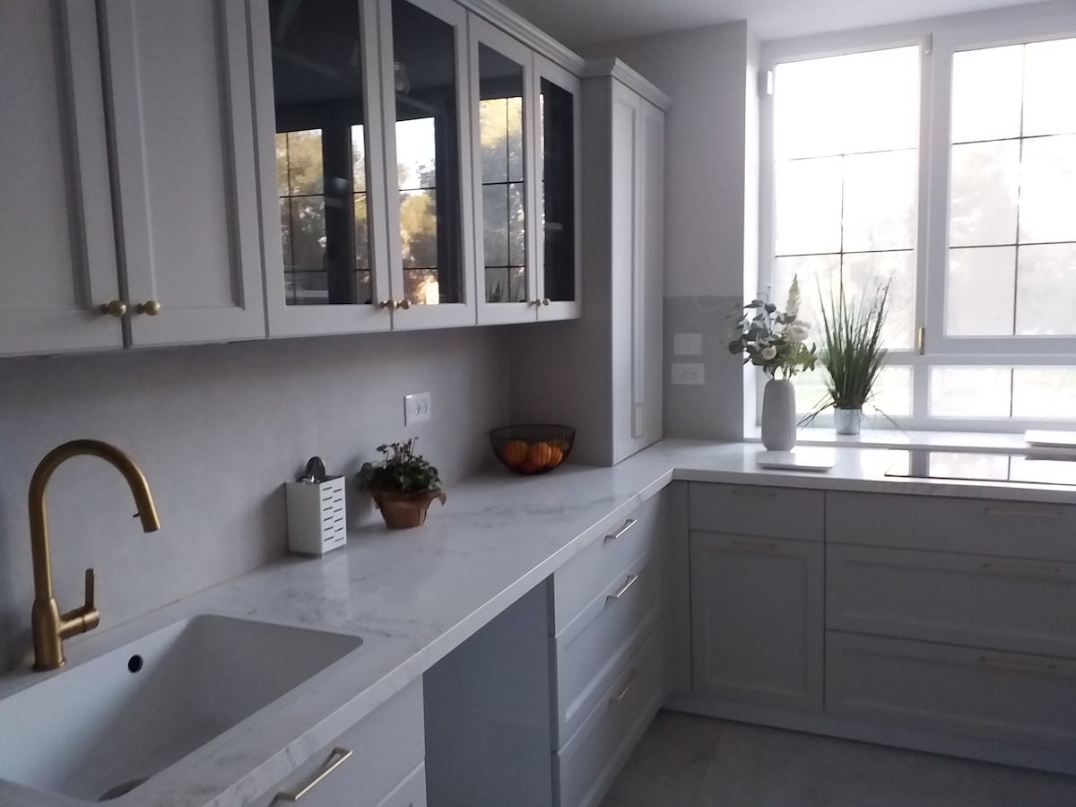 A modern kitchen features sleek white cabinetry with brass hardware. A large, white countertop extends alongside a window with a view. Simple decor includes potted plants and a fruit bowl, creating a clean and functional cooking space illuminated by natural light.