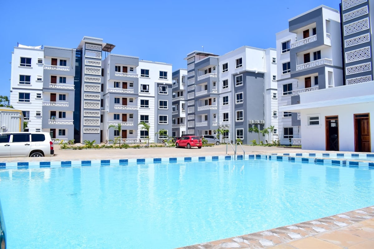 A large swimming pool is surrounded by modern apartment buildings with a mix of white and gray façades. The water is clear and inviting, reflecting the bright blue sky. Cars are parked nearby, contributing to the vibrant atmosphere of the area.