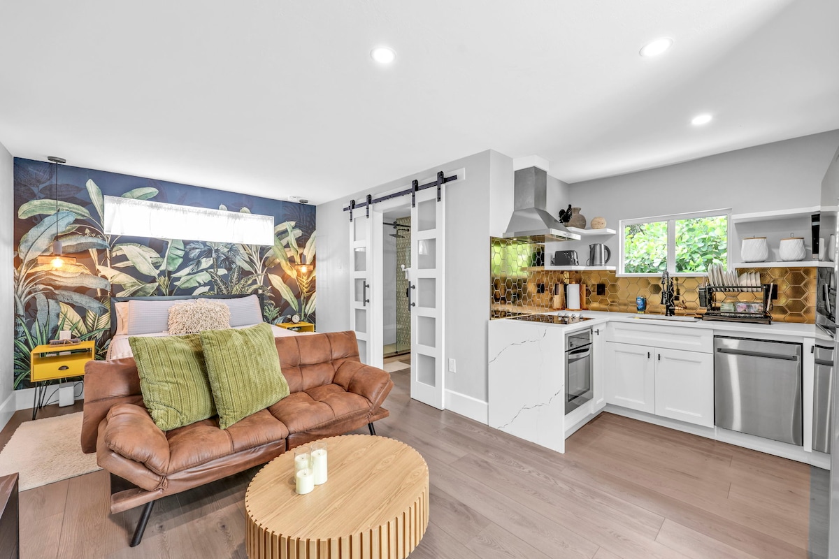 A modern open-concept living area features a comfortable brown leather sofa adorned with olive green cushions. A round wooden coffee table sits in the center. The kitchen showcases sleek white cabinetry and a stylish backsplash, with natural light illuminating the space through large windows.