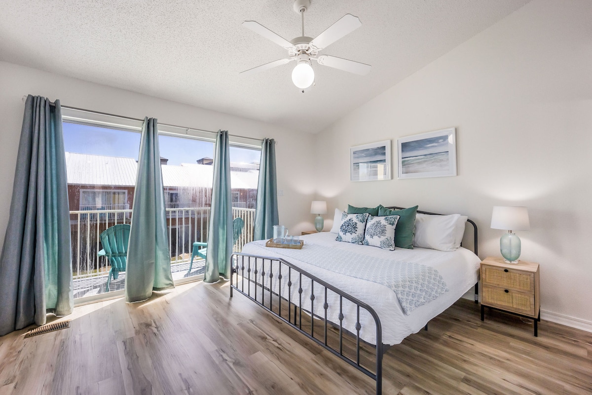 A bright bedroom is showcased with a spacious bed adorned with soft linens and decorative pillows. Light wood flooring complements the soothing color palette. Large glass doors lead to a balcony, allowing natural light to fill the space, while a ceiling fan contributes to air circulation.