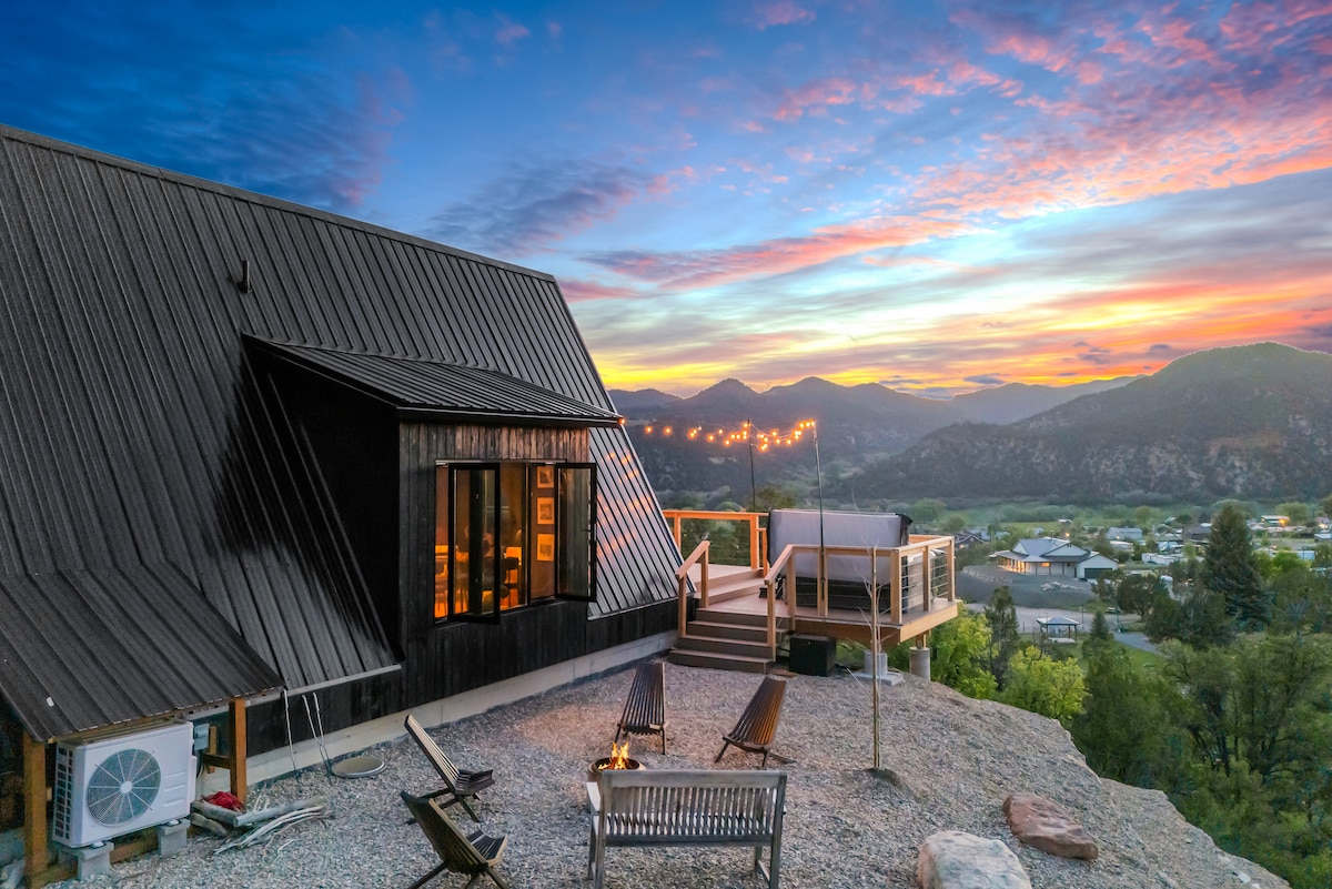 A modern A-frame cabin is depicted against a colorful sunset, surrounded by mountains. Outdoor seating is arranged near a fire pit, with a hot tub positioned on a deck. The cabin features large windows, allowing glimpses of the inviting interior, with soft lighting illuminating the space.