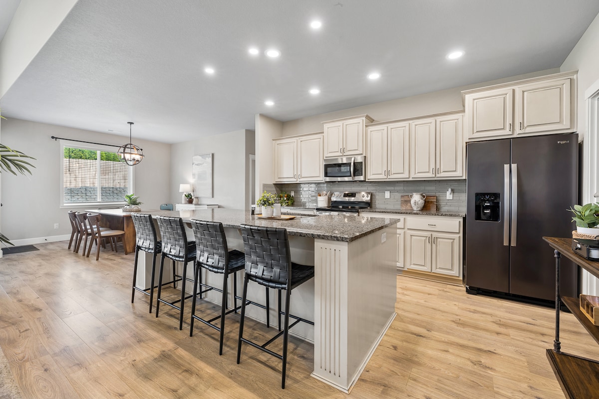 The kitchen features an island with seating for six, surrounded by light-colored cabinetry and layered with expansive countertops. A stainless steel refrigerator is positioned nearby, and a dining area with a wooden table is visible in the background. Natural light enters through a window, illuminating the space.