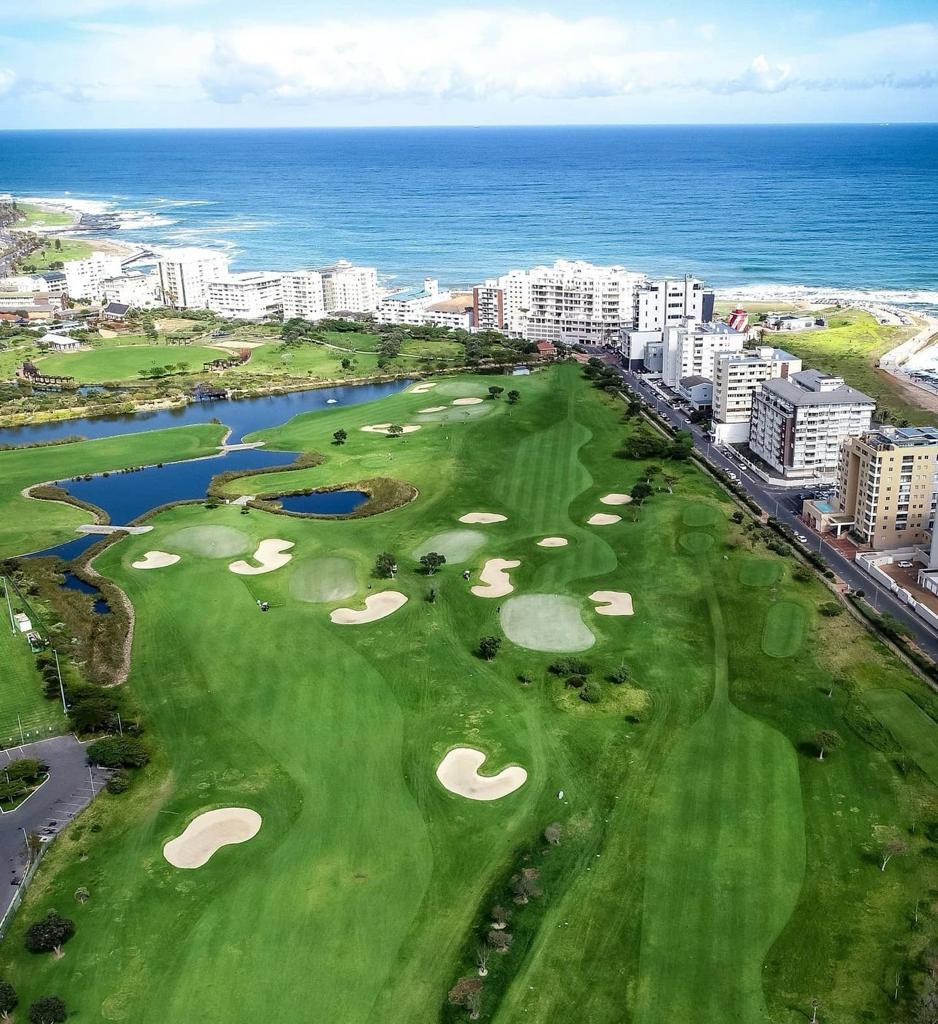 An aerial view captures the expansive green golf course, interspersed with sand traps and a serene pond. The coastline and the Atlantic Ocean stretch into the distance, with neatly arranged buildings lining the shore, creating a harmonious balance between nature and urban living.