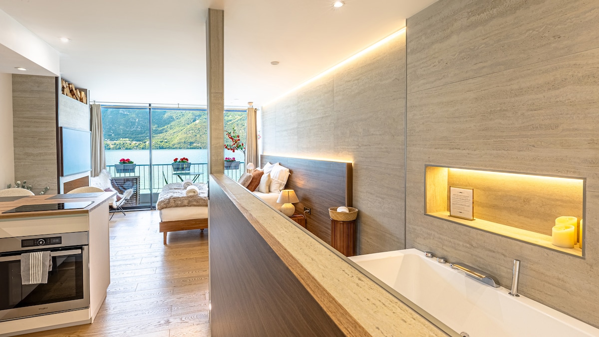 A modern open-plan living space showcases a spacious kitchenette with sleek cabinetry, a luxurious whirlpool bathtub, and a comfortable seating area. Large windows provide stunning views of the lake, allowing natural light to fill the room, while subtle lighting accents enhance the overall design.