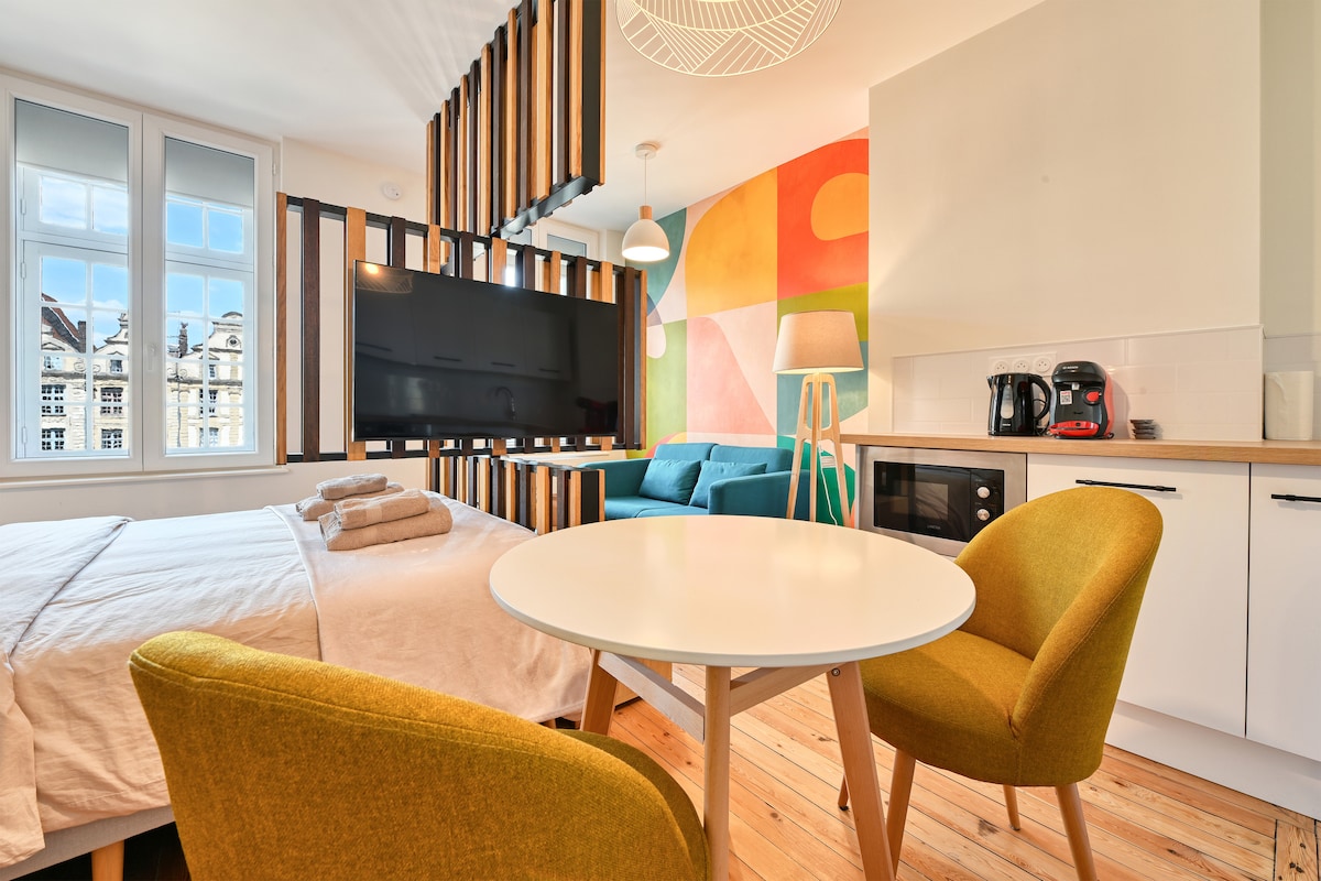 An inviting studio space features a spacious bed with neatly arranged towels. A television is mounted on a decorative wall, while a table is surrounded by two stylish chairs. A bright sofa and kitchenette are visible, complemented by colorful artwork on the walls.