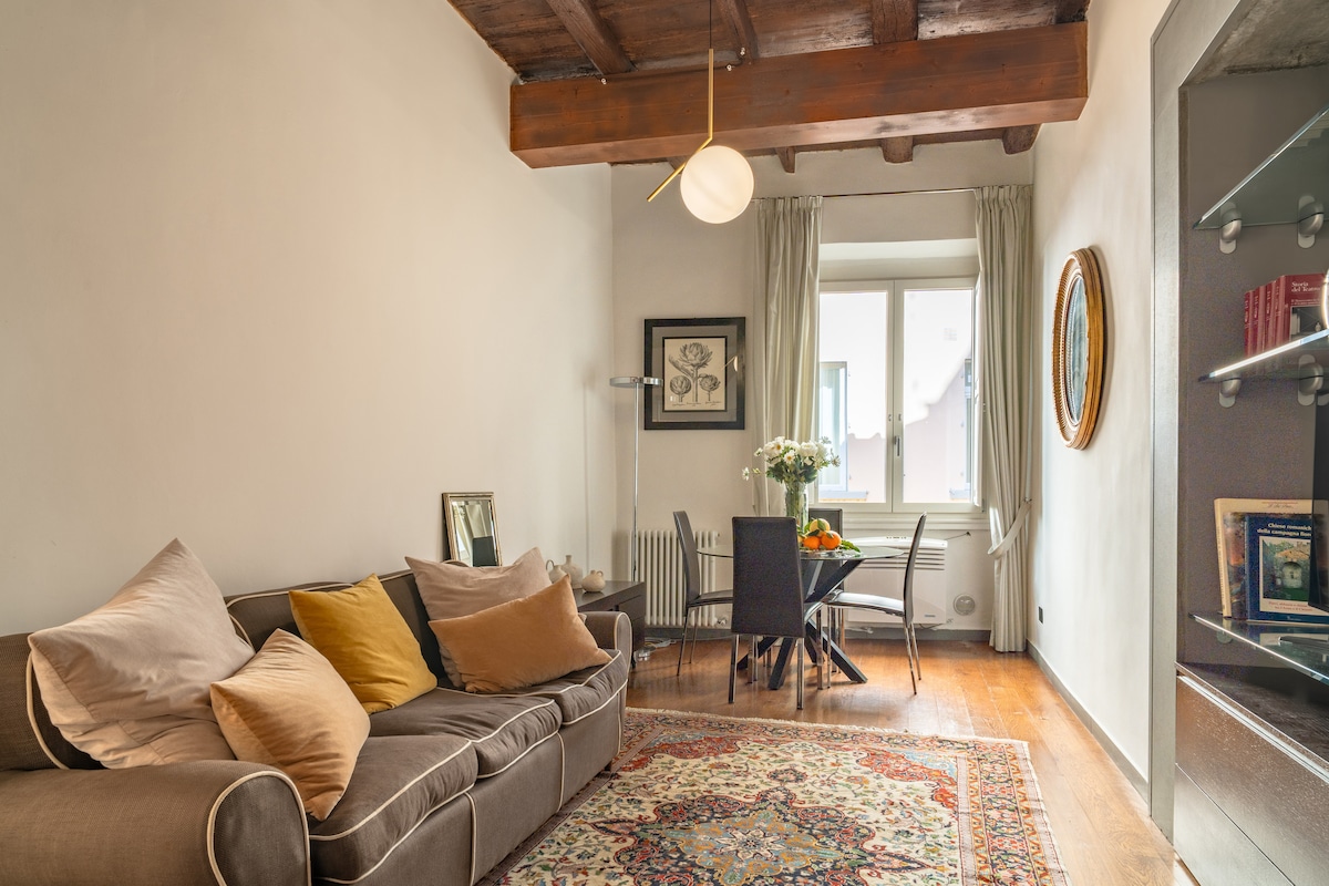 A living area is seen with a comfortable sofa adorned with decorative pillows. A dining table is positioned nearby under a large window, complemented by light-colored curtains that gently frame the view. Warm wood beams and a patterned rug enhance the inviting atmosphere.