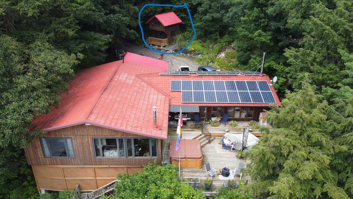 Aerial view captured from above showcases the property, featuring a main cottage with solar panels on a red roof and a secondary wooden bunkhouse nestled among lush greenery. The outdoor deck extends from the main building, surrounded by trees that provide a serene setting.