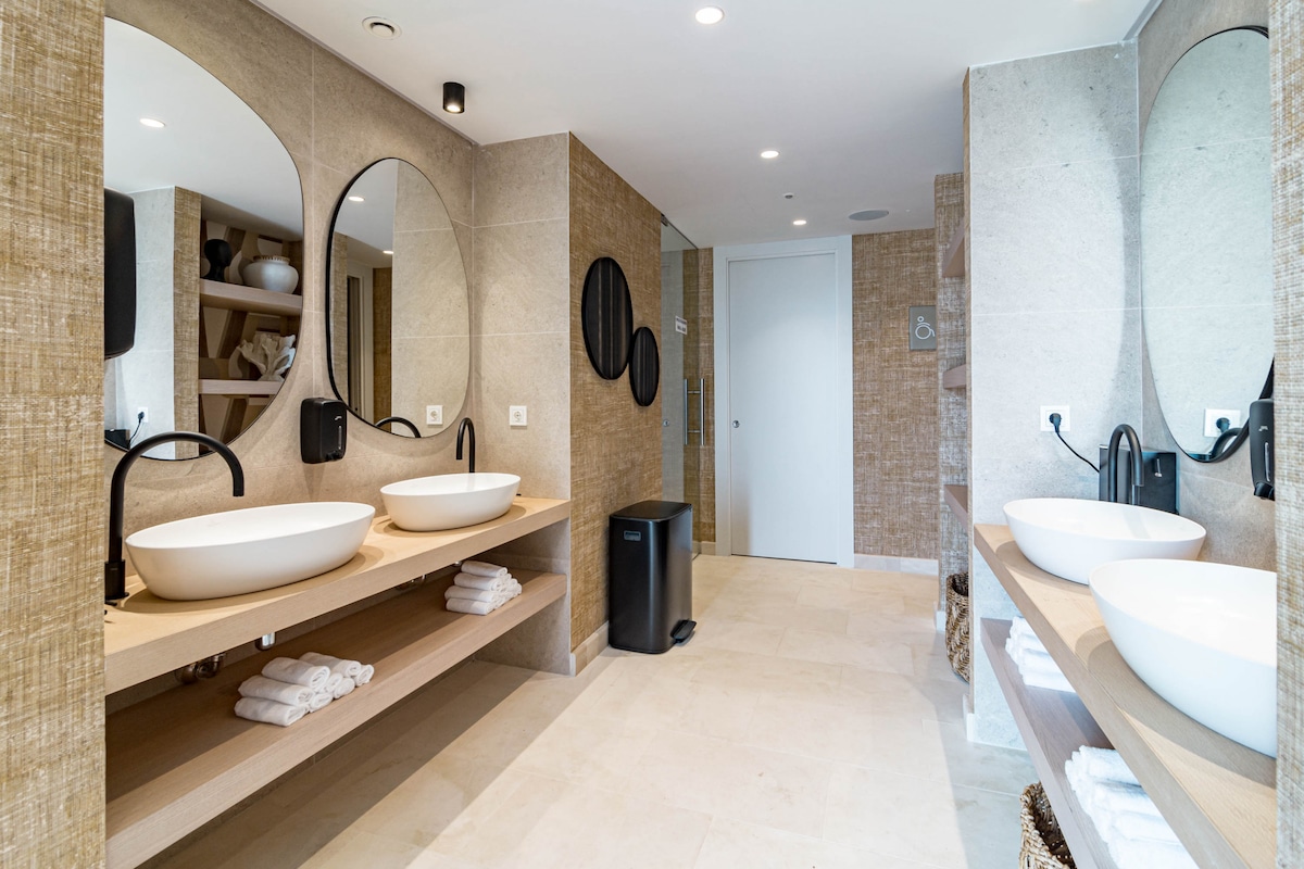 A modern bathroom design features dual vessel sinks on a wooden vanity, with large round mirrors positioned above. Soft lighting enhances the neutral tones of the tiled walls. Shelving displays decorative items, while neatly stacked towels are arranged below. A sleek black wastebin complements the overall aesthetic.