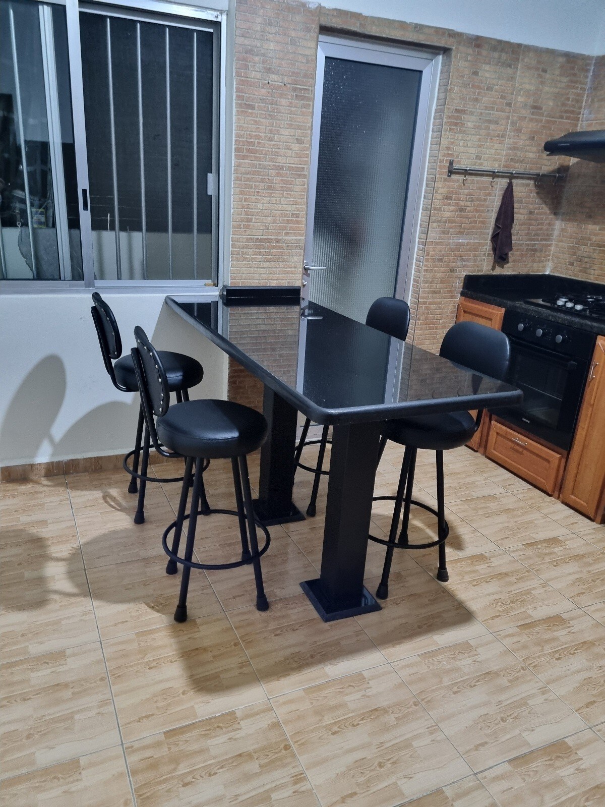 A modern kitchen area features a sleek black countertop with four high-backed chairs. Natural light enters through a large window, revealing a stainless steel stove and wooden cabinetry. The floor is tiled in neutral tones, enhancing the clean and functional space.