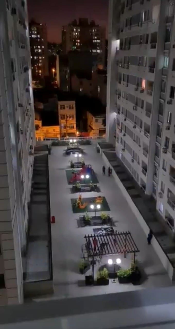 An aerial view captures the well-lit courtyard of a residential complex, featuring colorful play structures and seating areas. Surrounding buildings are visible, with warm lights creating a lively evening atmosphere. Pathways and greenery add to the inviting design of the community space.