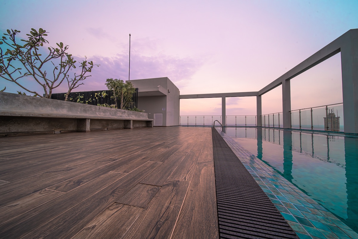 The rooftop pool area is showcased with modern tile accents and wooden decking. A few green plants provide natural touches, while a striking skyline can be seen in the background. The space offers a sense of openness and tranquility, highlighted by the soft evening light.