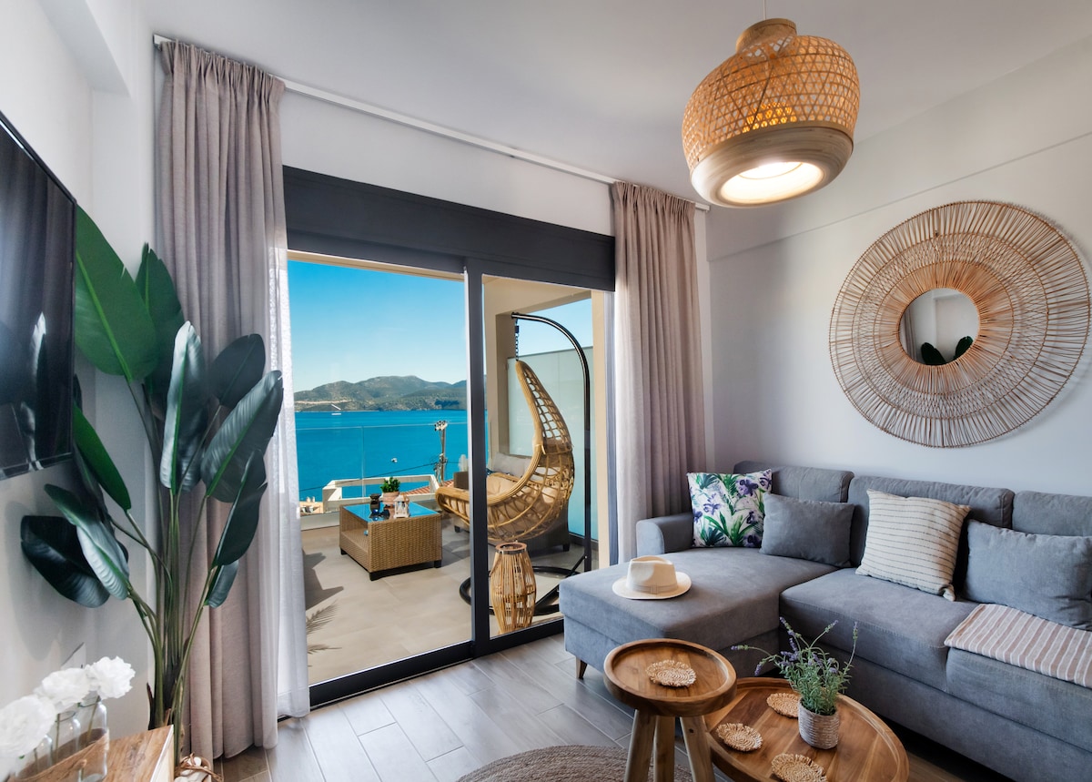 A stylish living area features a gray sectional sofa adorned with decorative pillows. A large mirror and a circular basket light fixture add interest, while sliding glass doors reveal a scenic view of the sea and outdoor seating on the patio.