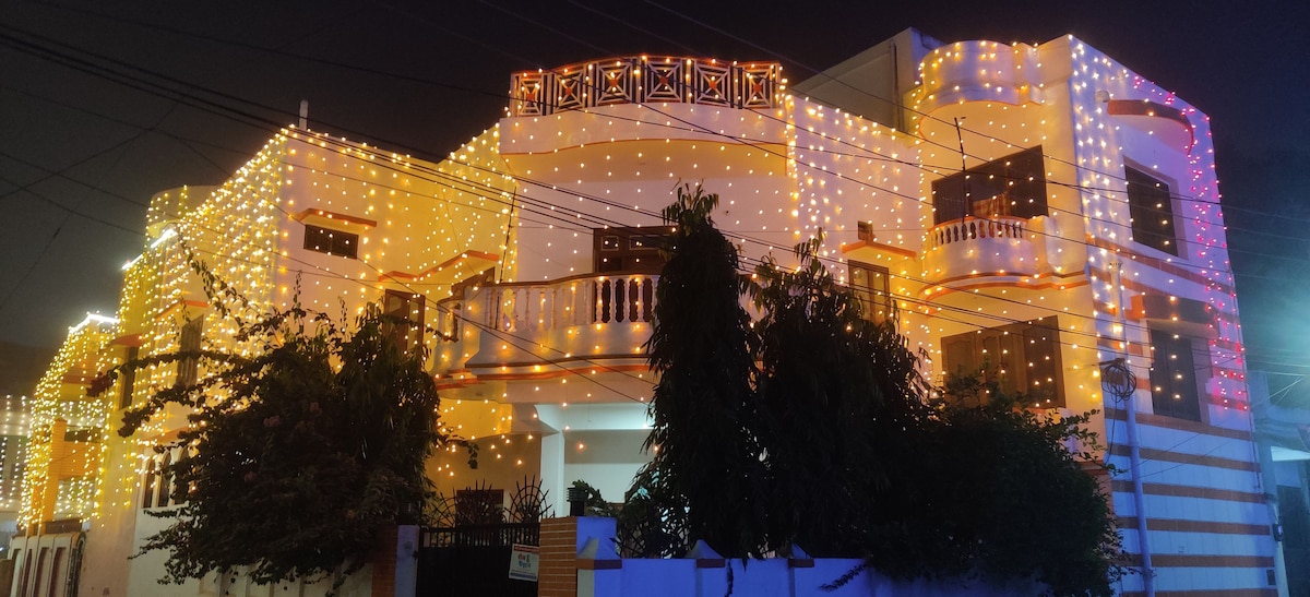 The exterior of the building is adorned with numerous decorative lights, creating a festive ambiance. The structure features multiple balconies and windows, surrounded by lush greenery. The night sky enhances the vibrant colors of the lights against the white walls.