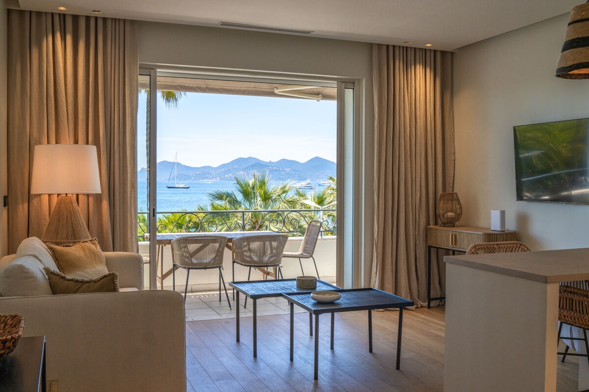 A bright living area features a comfortable sofa facing a large window with a panoramic view of the sea. A small dining table with chairs is positioned on the balcony, with soft curtains framing the view and natural light filling the space.