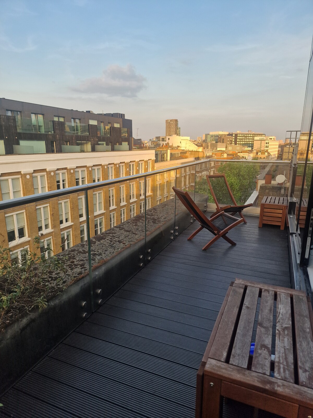 A private balcony features wooden decking and glass railing, offering views of the surrounding buildings. Two lounge chairs are positioned for relaxation, alongside a wooden bench. The skyline is visible, illuminated by soft evening light.