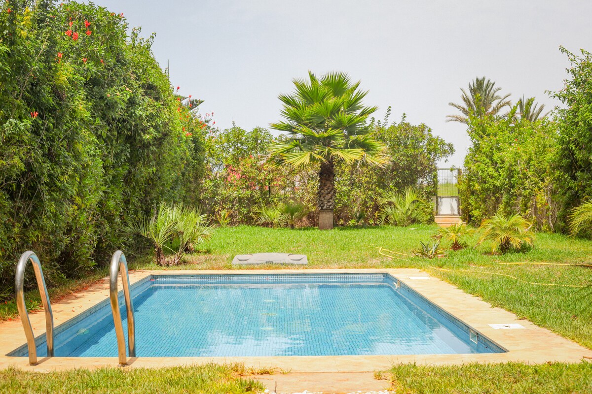A private swimming pool is framed by lush greenery and provides a refreshing retreat. Surrounding palm trees and flowering plants enhance the natural setting, while a sunbathing area with a lounge chair is visible along the edge of the pool.