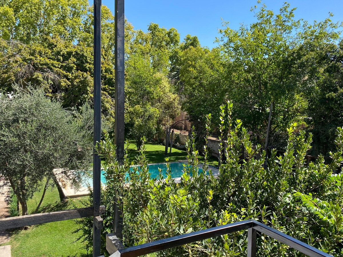 A view from the balcony showcases a lush garden with tall trees and well-maintained greenery. In the distance, a serene swimming pool is visible, inviting relaxation and tranquility within the warm, sunlit landscape.